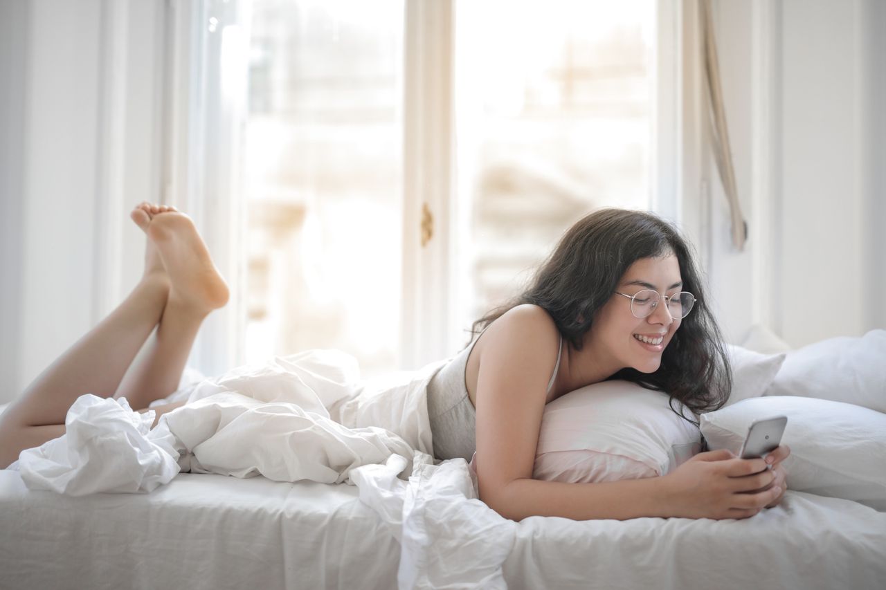 faq-img Smiling woman in a cozy bedroom, using smartphone, embracing relaxation and comfort.